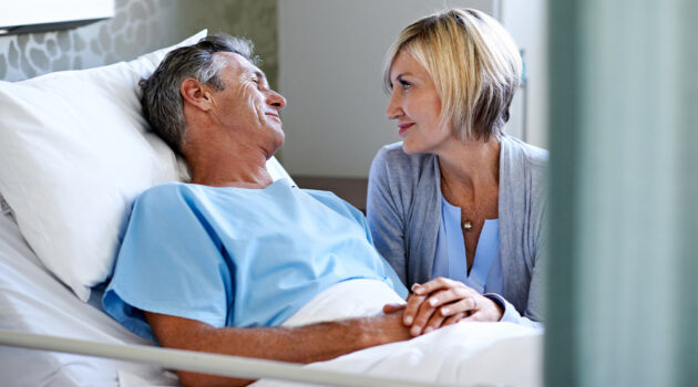 Man in bed, woman standing next to him looking at him.