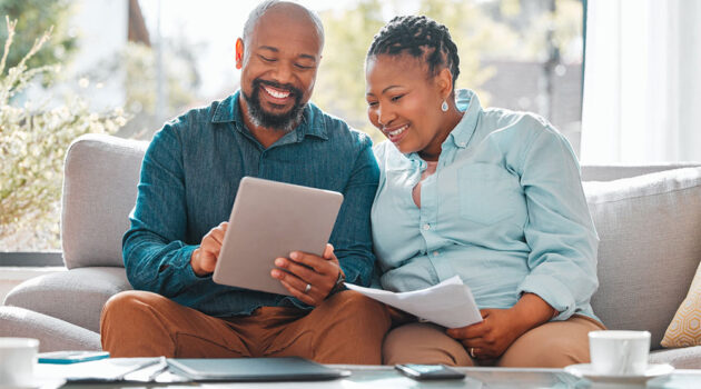 Mature senior couple sits on sofa and uses the internet on a tablet.